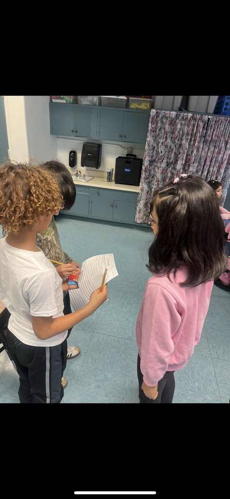 Three students stand in a cluster in the middle of a classroom, collaborating on a survey. One student in a white t-shirt holds a worksheet while another student points to it with a pencil. A blue cabinet and a patterned curtain are visible in the background.