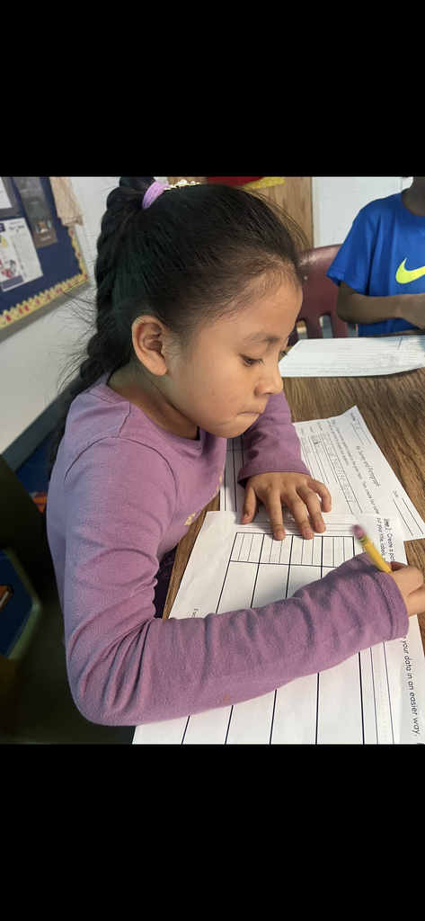 A girl with a long dark braid, wearing a purple long-sleeved shirt, concentrates as she draws on a pictograph worksheet. She is leaning over a wooden table in a bright classroom, with another student in a blue Nike shirt partially visible next to her.