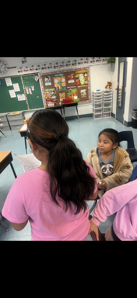 A student with long dark hair in a pink shirt (viewed from behind) stands and talks to a seated classmate wearing a panda t-shirt and a fur-lined jacket. They appear to be conducting a survey. The classroom walls are decorated with colorful "How to Calm Down" and "Problem-Solving Steps" posters.