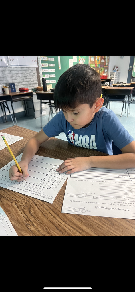 A young boy in a blue "NBA" t-shirt sits at a desk, carefully drawing symbols onto a "Favorite Chips" pictograph. Two worksheets are on the desk: one for collecting tally marks and one for the final pictograph. He is highly focused on his work.