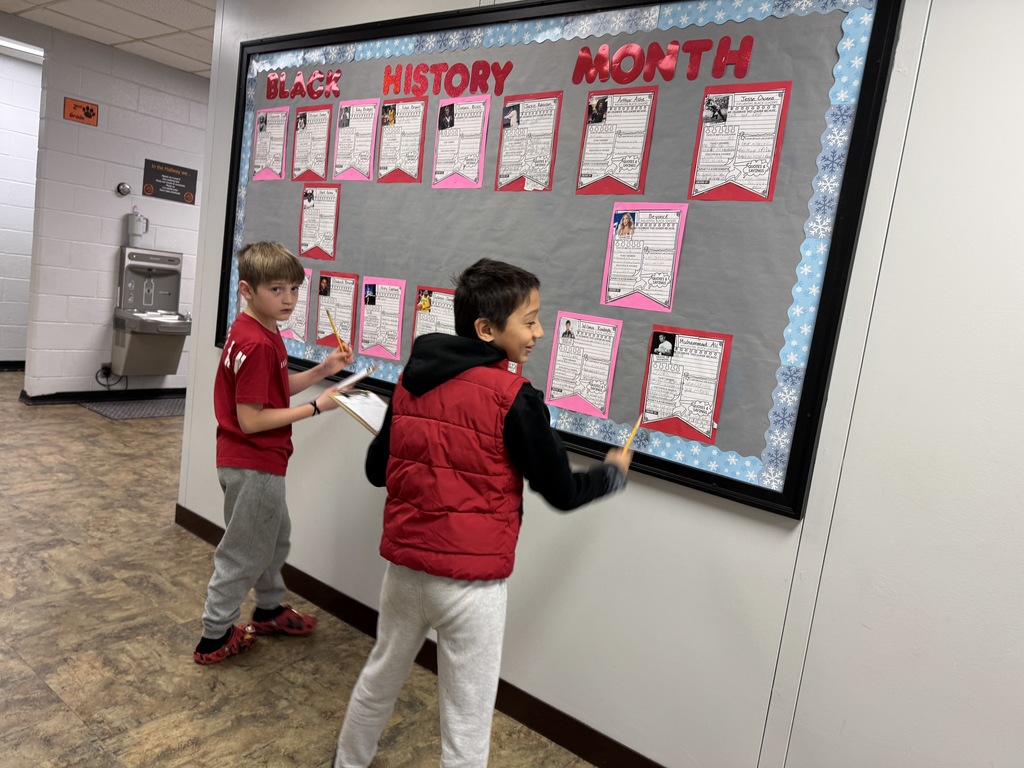 Two elementary students stand in a school hallway reading and writing on a Black History Month bulletin board featuring student-created profiles of influential Black leaders, with papers displayed on a gray background and red lettering.