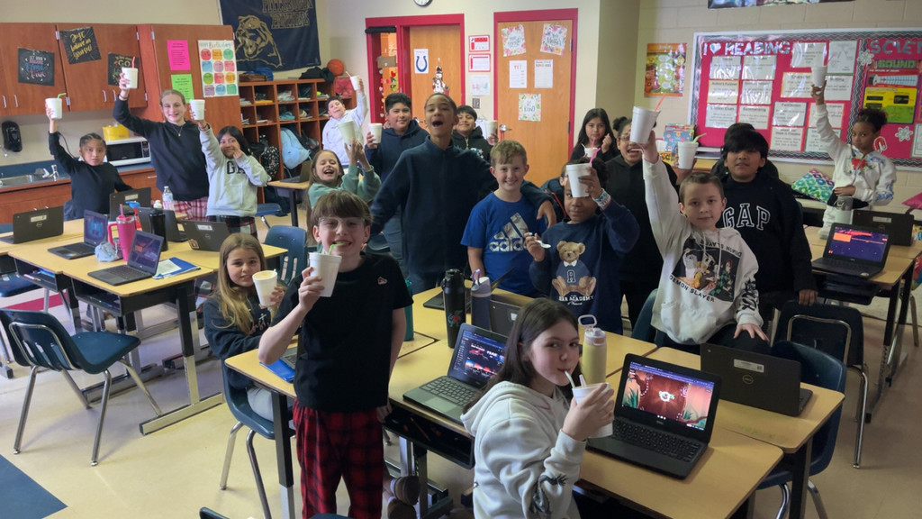 A classroom full of students wearing pajamas raise white cups in the air while smiling and cheering during a reward celebration. Several Chromebooks sit open on desks throughout the room.  Image 3: