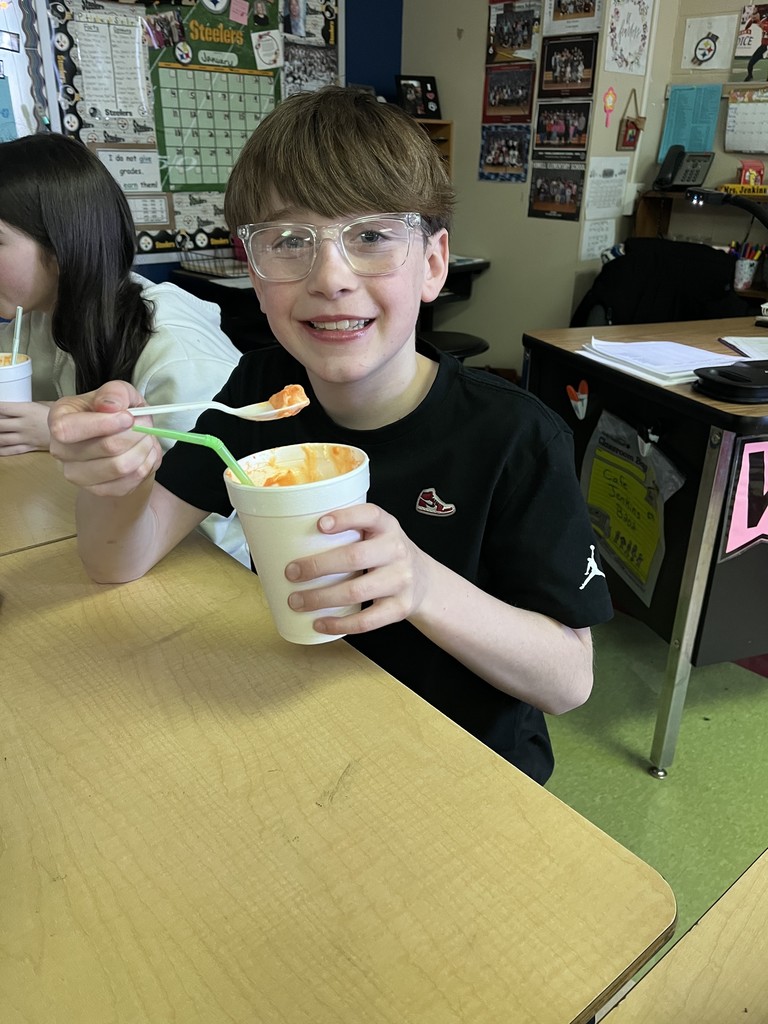 A student wearing glasses smiles while holding a white cup and spoon filled with orange-colored float. A classroom desk and teacher workspace are visible behind them.
