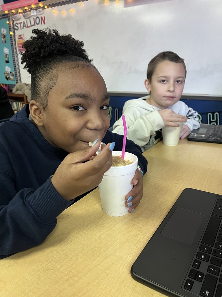 A student sits at a desk eating a float with a spoon from a white cup while smiling. Another student sits beside them holding a cup, with Chromebooks visible on the desks.
