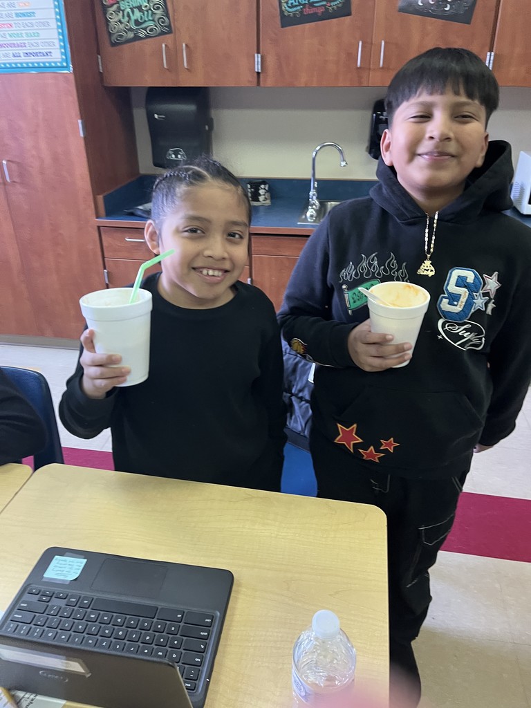 Two elementary students stand in a classroom smiling and holding white cups with straws, enjoying float treats during a pajama-themed classroom reward celebration. A desk with a Chromebook is visible in the foreground.