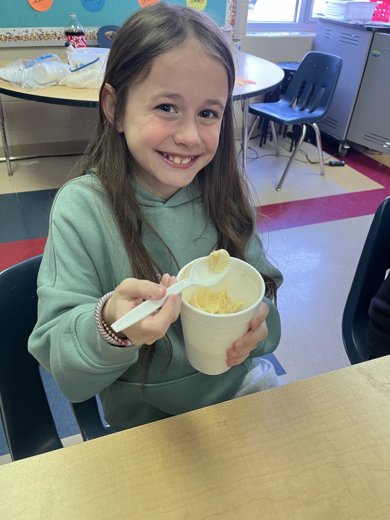 A smiling student sits at a desk holding a spoonful of float from a white cup. A classroom table and chairs are visible in the background.