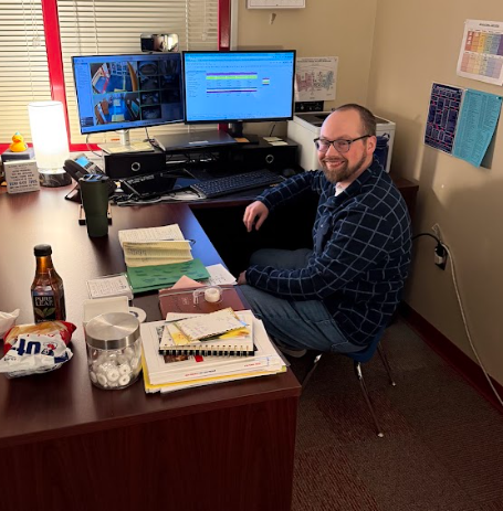 Alt text: School administrator sitting at a desk in a small office using two computer monitors, smiling while seated on a small student chair as part of a classroom reward challenge.