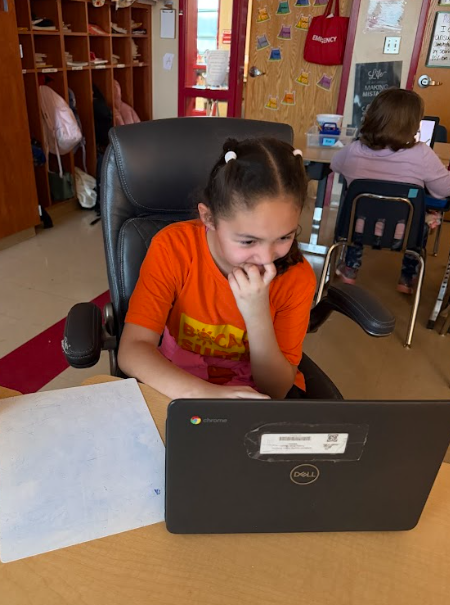 Alt text: Elementary student wearing an orange shirt sitting in a black office chair while working on a Chromebook at a classroom desk.