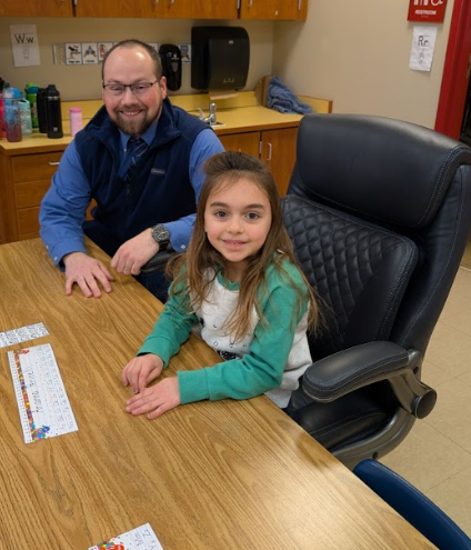 Alt text: Elementary student sitting in a large black office chair at a classroom table with a school administrator standing behind her, both smiling during the attendance reward activity.