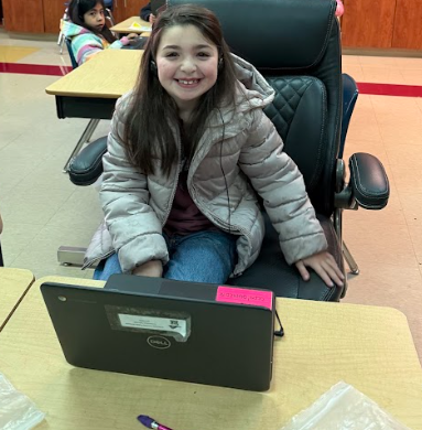 Alt text: Elementary student wearing a winter jacket sitting in a black office chair at a desk with a Chromebook open, smiling during the attendance challenge reward activity.