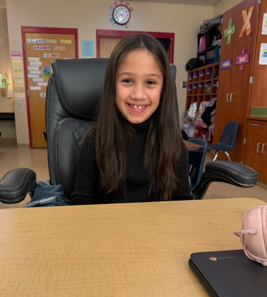 Alt text: Elementary student smiling while sitting in a large black office chair at a classroom table during the attendance reward celebration.