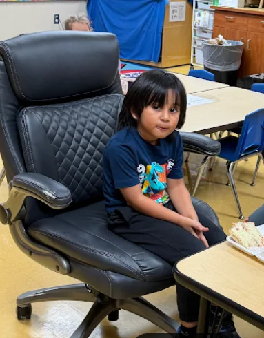 Elementary student sitting in a large black office chair at a classroom table, looking toward the camera while holding a snack plate during the attendance reward activity.