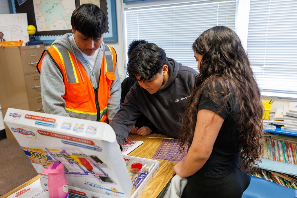 A CTEC electrical program student wearing an orange safety vest stands with two students at a classroom table as they work together on a Snap Circuits electronics project.