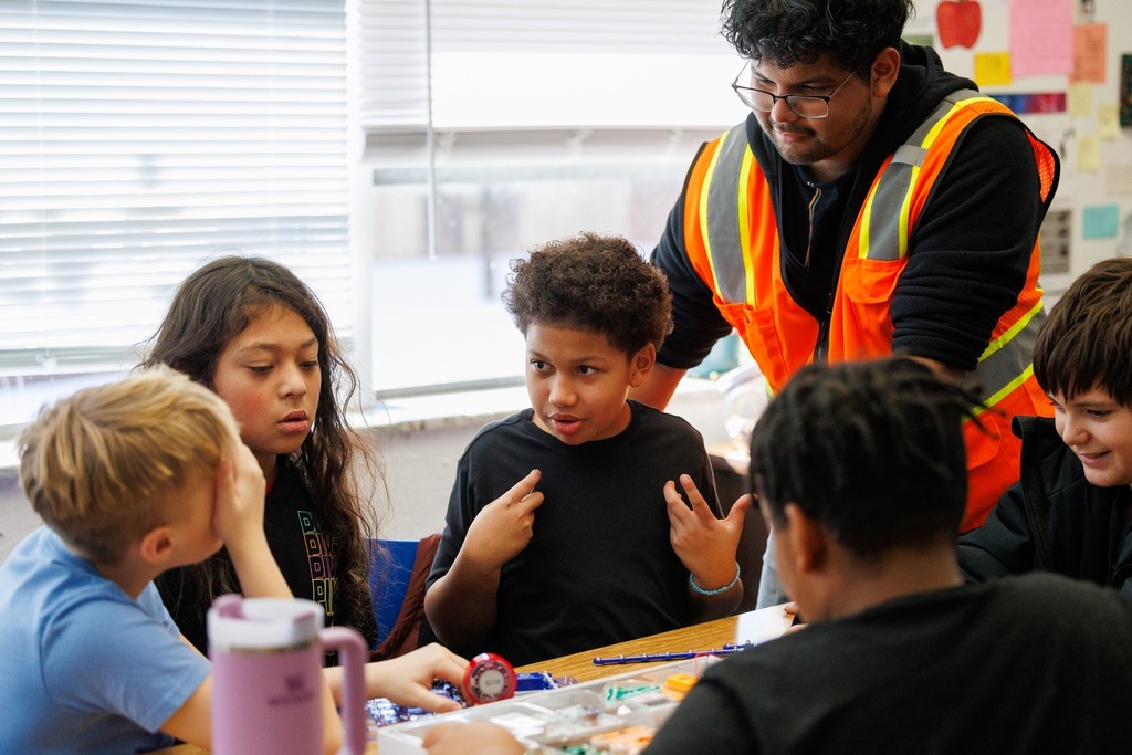 A CTEC electrical program student wearing an orange safety vest leans over a table while elementary students work together on an electronics activity, with building materials and tools spread out in a classroom.
