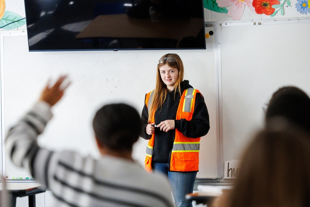 A CTEC electrical student wearing a safety vest stands at the front of a classroom, speaking to younger students as one raises a hand.