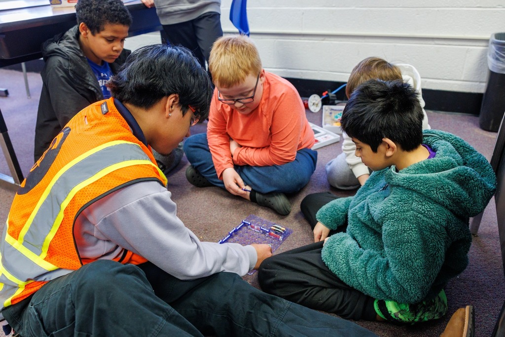 A CTEC electrical student wearing a safety vest sits on the floor with elementary students, helping them build a Snap Circuits project.