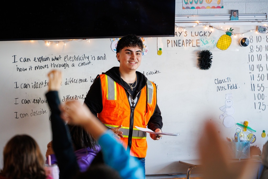 A CTEC electrical program student wearing an orange safety vest stands at the front of an elementary classroom holding papers, while children raise their hands in the foreground. A whiteboard behind him shows handwritten notes about electricity and classroom decorations.