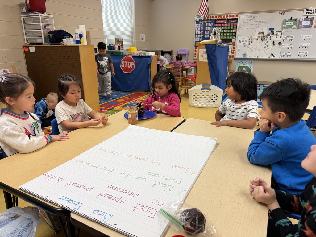 Several young students sit around a classroom table watching a classmate spread peanut butter and birdseed onto a pinecone on a plate. A large chart with step-by-step directions for making a pinecone feeder lies on the table, and classroom centers and play areas are visible behind them.