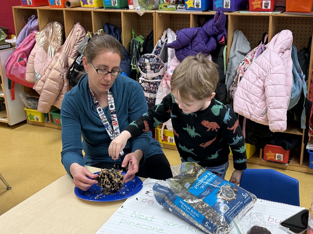 A teacher and a young student work together at a classroom table to coat a pinecone with birdseed on a blue plate. A large bag of birdseed and a chart with written steps are on the table, and cubbies filled with coats and backpacks line the wall behind them.