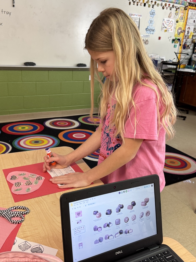 Student standing at a desk gluing a fraction label onto a red Valentine chocolate box project. A Chromebook on the desk shows a slide with different chocolate types. Cut paper hearts and craft supplies are nearby.