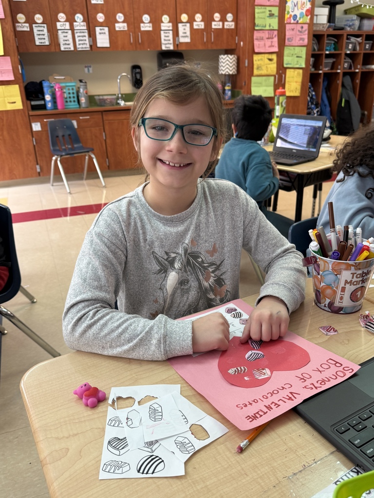Student wearing glasses sits at a desk placing small chocolate cutouts onto a pink heart paper for a Valentine fraction activity. Markers, a pencil, and printed chocolate templates are spread on the desk, with classroom cabinets and posters in the background.