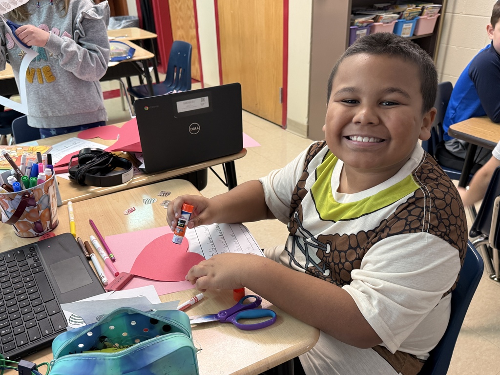 Smiling student seated at a classroom desk working on a Valentine chocolate fraction craft. The student holds a glue stick over a pink paper heart with small chocolate cutouts. A Chromebook, scissors, markers, and pencil pouch are on the desk.