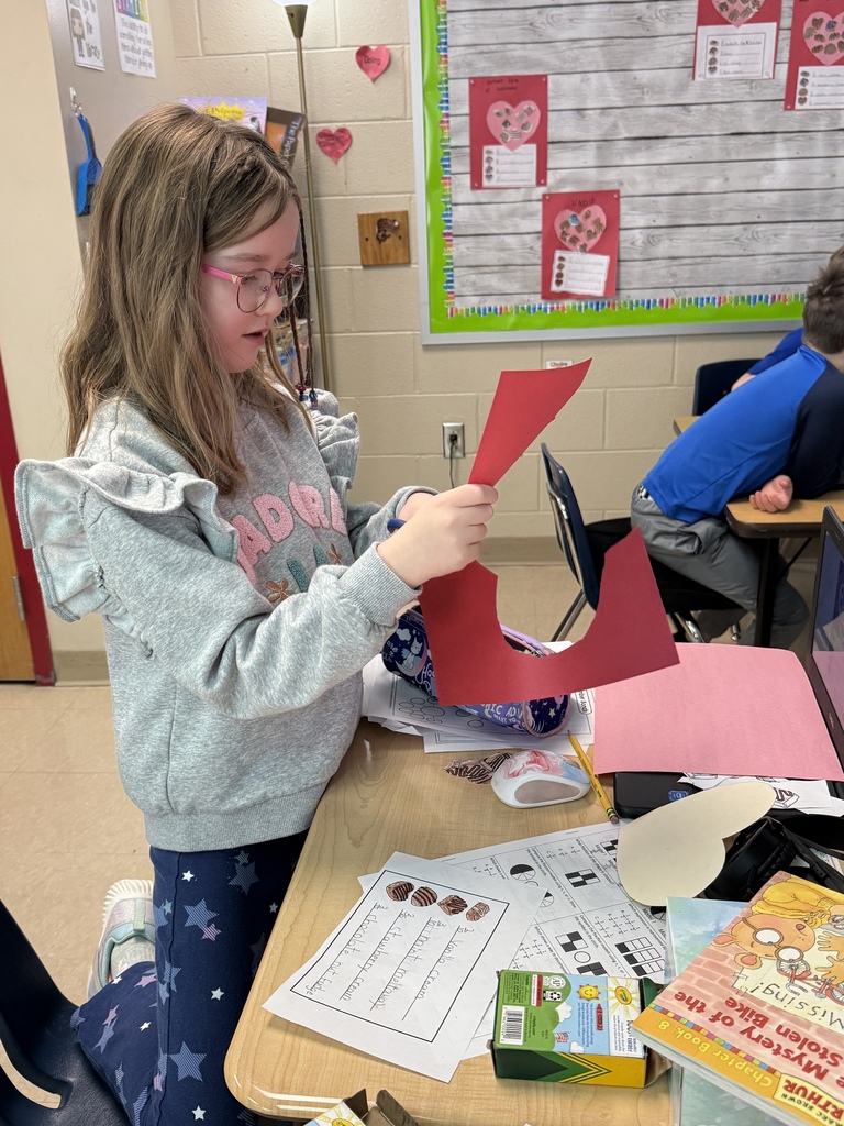 Student cutting a large red paper heart at a classroom desk during a Valentine fraction craft activity. Worksheets, crayons, books, and a Chromebook are visible on the table, with a Valentine project bulletin board in the background.