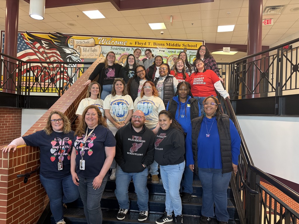 A group of middle school staff members stand together on a staircase inside Floyd T. Binns Middle School. Many are wearing matching or coordinated outfits for a themed spirit day, smiling at the camera. A colorful school mural reading “Welcome to Floyd T. Binns Middle School” is visible in the background.
