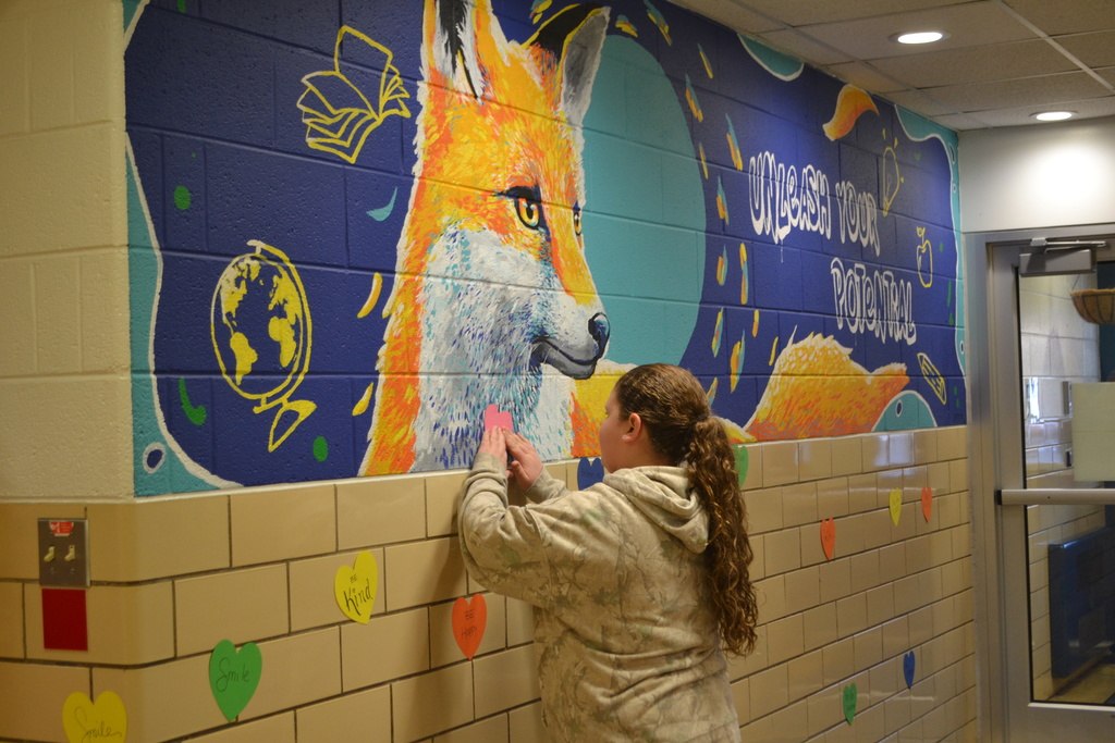 Student is placing a heart on the school wall by the school mural- unleash your potential.  There are other colored hearts on the wall too.