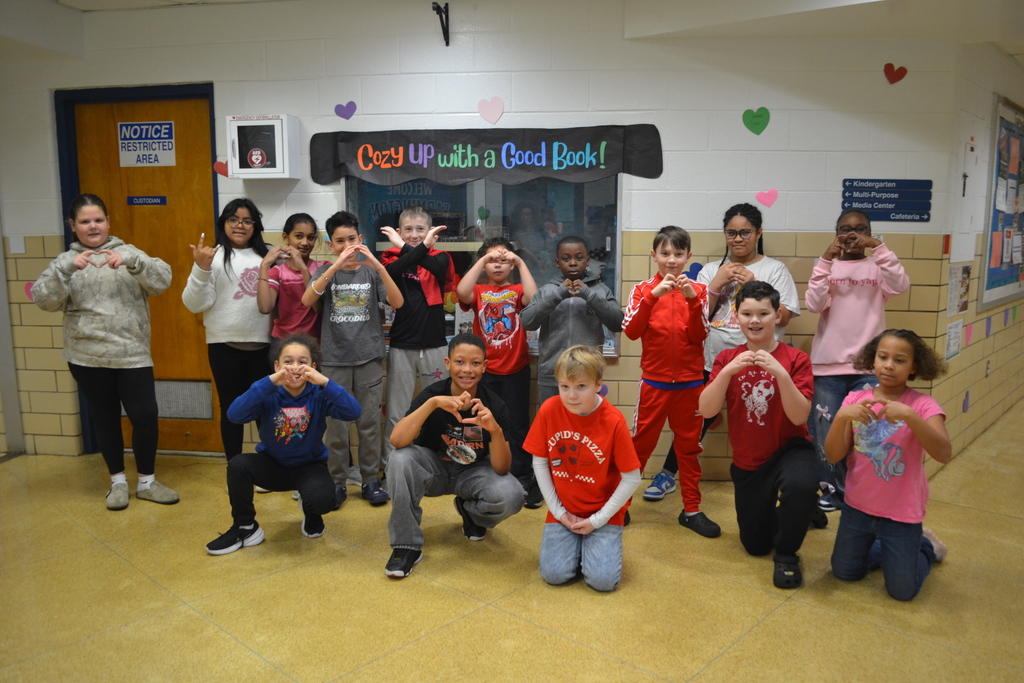 A group of elementary students pose in a school hallway decorated with colorful paper hearts and inspirational messages for Kindness Week. The students are smiling and making heart shapes with their hands. A sign above a display case reads, “Cozy Up with a Good Book!”