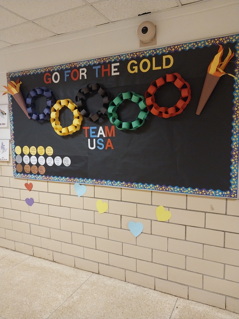 Hallway bulletin board with a black background displaying the message “Go for the Gold” and “Team USA.” The board features five Olympic rings made from colored paper chains in blue, yellow, black, green, and red, along with paper torch decorations on both sides. Paper circles on the left resemble medals and include student names or events, and colorful heart cutouts are displayed on the wall below the board.