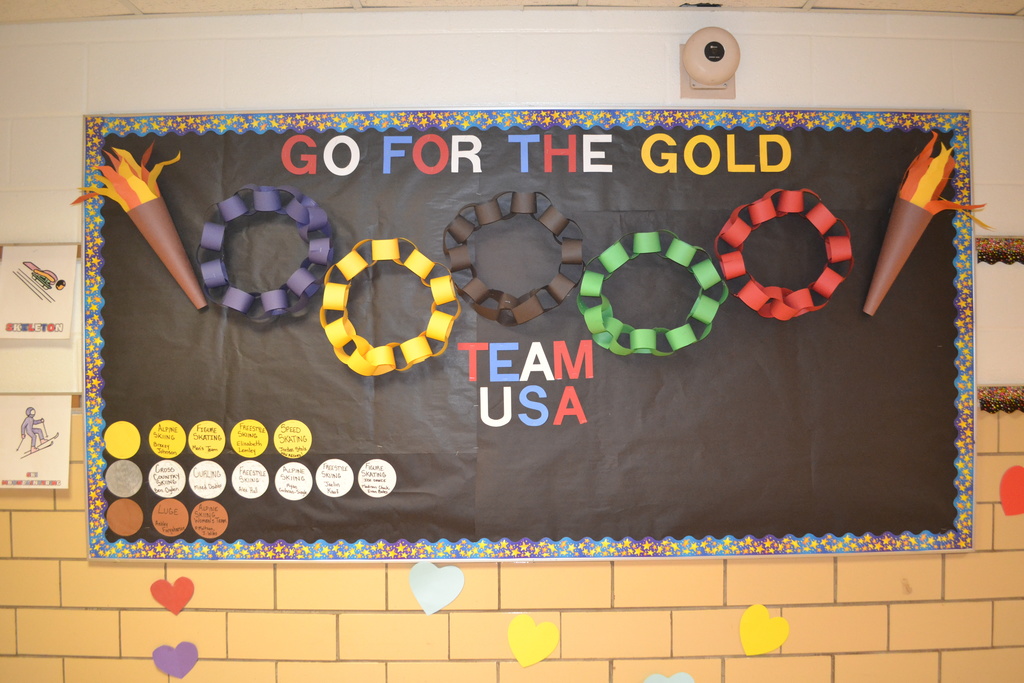 Hallway bulletin board with a black background displaying the message “Go for the Gold” and “Team USA.” The board features five Olympic rings made from colored paper chains in blue, yellow, black, green, and red, along with paper torch decorations on both sides. Paper circles on the left resemble medals and include student names or events, and colorful heart cutouts are displayed on the wall below the board.