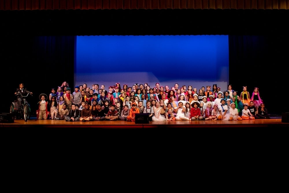 Wide stage photo of a large elementary school musical cast gathered together for a group picture under blue stage lighting, with dozens of students in colorful costumes seated and standing across the stage.