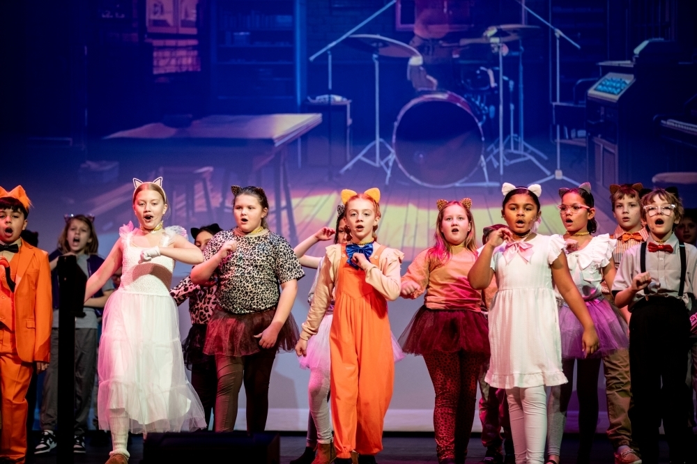 Row of student performers wearing animal ears and themed costumes singing and doing choreography at the front of the stage, with a band set and props visible in the background.