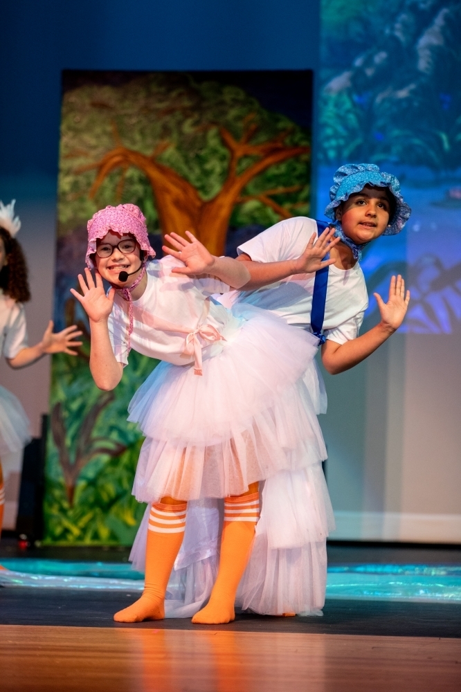Two student performers in matching white costumes with pastel accessories and hats pose back-to-back mid-dance, smiling and holding their hands up as part of a choreographed number.