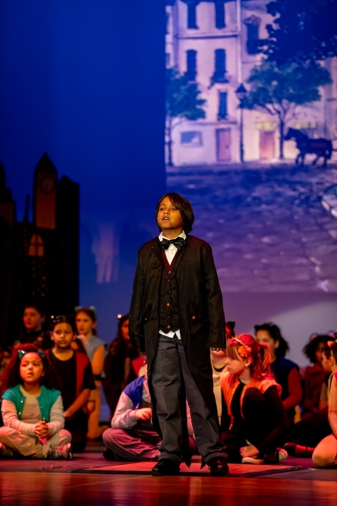 Student soloist in a dark coat and bow tie stands center stage singing, with other costumed students seated behind as the ensemble, against a city street backdrop.
