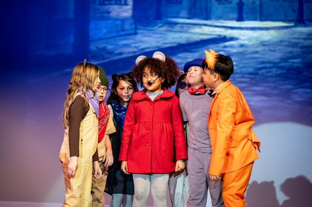 Small group of student performers in costume standing close together on stage, including animal-themed outfits and bright colors, gathered around a central character in a red coat during a musical scene.