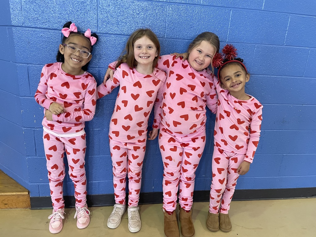 4 girls pose in the school gym wearing matching pajamas (pink with red hearts). The wall is blue.