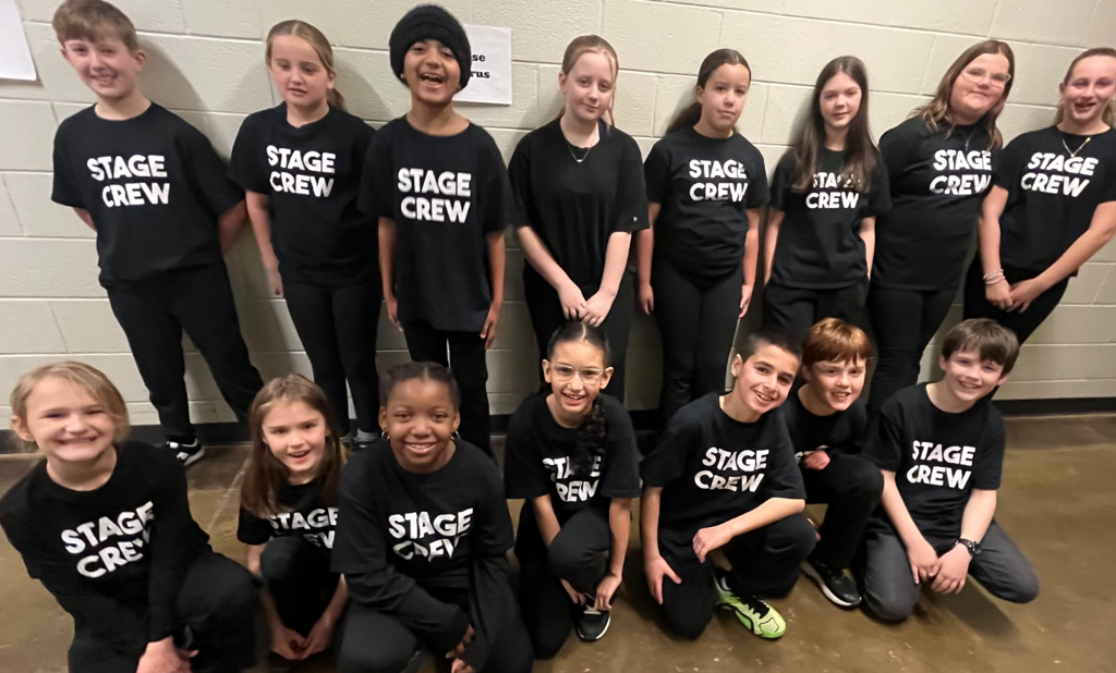 A group of stage crew members pose together in a school hallway. They wear matching black shirts with “STAGE CREW” printed in bold white letters. The students smile proudly for the photo.