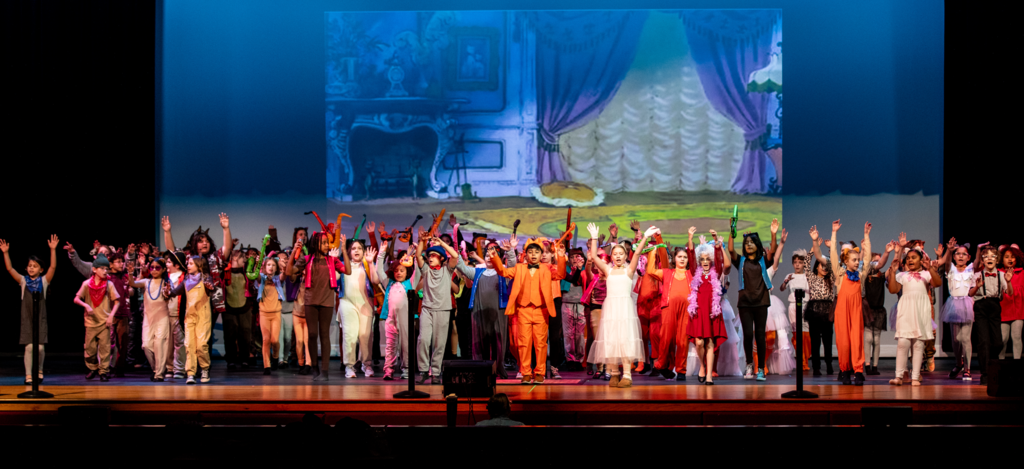 A wide stage shot shows a large group of elementary students performing Aristocats Kids. The cast fills the stage with arms raised in celebration. Students wear colorful costumes including cat ears, bright dresses, vests, bandanas, and character outfits. A Parisian-style backdrop with curtains and a fireplace is projected behind them.