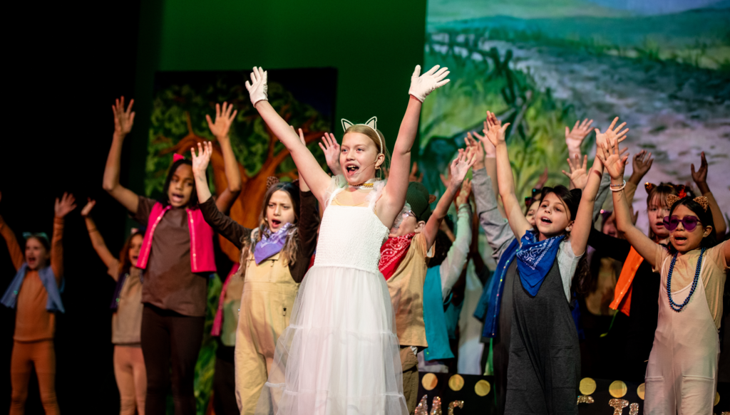 A student in a white dress with cat ears and white gloves stands center stage with arms raised confidently. Other students in coordinated costumes with bandanas and vests mirror the pose behind them. A painted countryside scene serves as the backdrop.