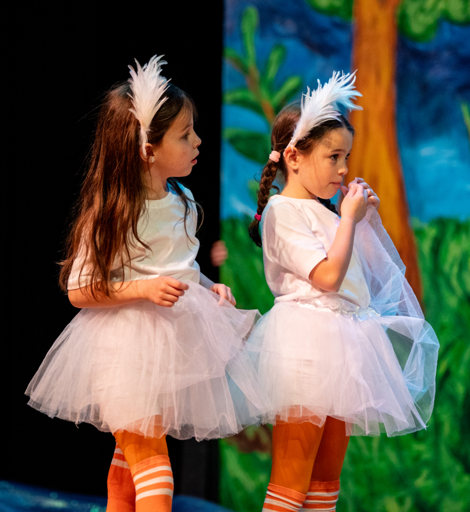 Two young students dressed in white ballet-style costumes with tulle skirts and white feather headpieces stand on stage. They wear orange-and-white striped tights and appear focused during a scene set against a painted outdoor backdrop with trees and greenery.