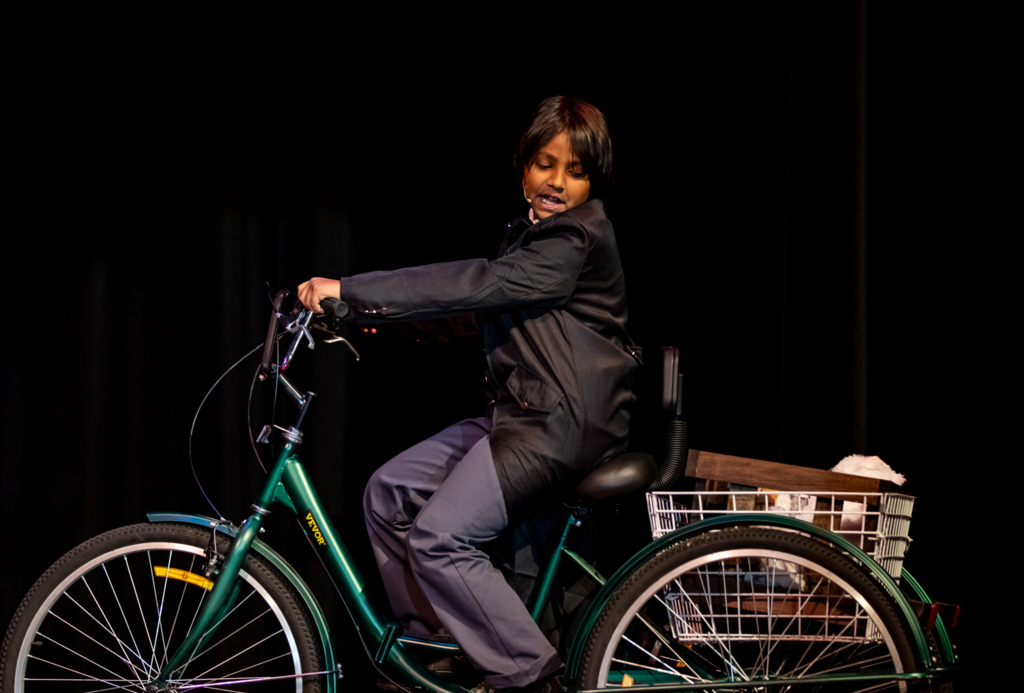 A student dressed in a black suit rides a green tricycle across the stage. A basket attached to the back of the tricycle contains props. The dark stage lighting highlights the character in motion.