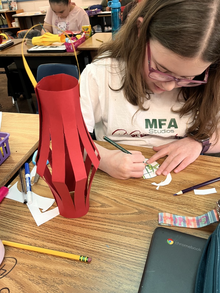 A student sits at a desk in a classroom drawing and coloring a small paper craft with a pencil. A completed red paper lantern stands upright on the desk beside markers, pencils, and a Chromebook, showing the Lunar New Year lantern-making activity in progress.