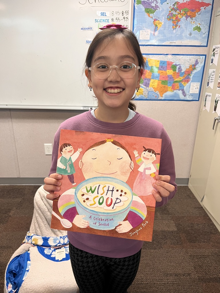 A student stands in a classroom smiling and holding the picture book Wish Soup: A Celebration of Seollal facing the camera. A classroom whiteboard and maps of the world and the United States are visible in the background.