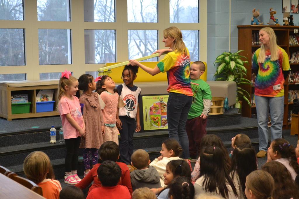 A presenter uses a large model toothbrush above a group of smiling students participating in the demonstration. Another staff member stands nearby as classmates watch from the floor.