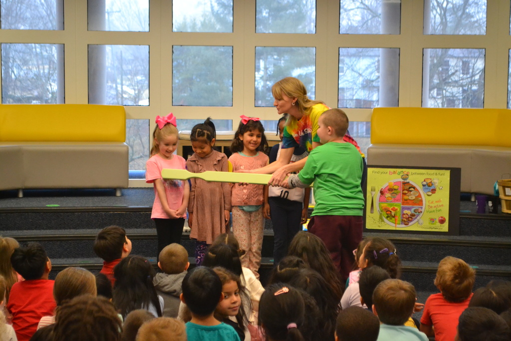 A presenter demonstrates with an oversized toothbrush prop while several young students stand beside her at the front of a library. Other students sit on the floor watching the interactive dental health presentation.
