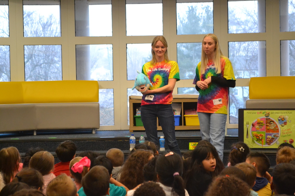 Two presenters wearing colorful tie-dye shirts stand at the front of a school library speaking to a large group of elementary students seated on the floor. One presenter holds a plush shark while discussing dental health.