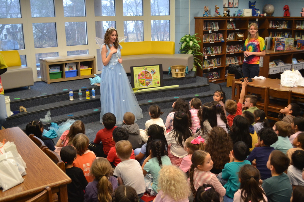 A person dressed as the Tooth Fairy in a light blue gown with wings stands at the front of a school library speaking to a seated group of elementary students during a dental health presentation. A staff member stands nearby.