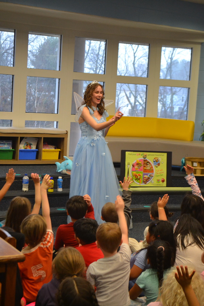 The Tooth Fairy gestures and smiles while speaking to students who raise their hands to answer questions during a dental health program in the school library.